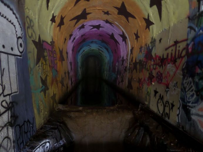 Flash photo of a graffitied tunnel with water covering the floor leading to a hole near the viewer.