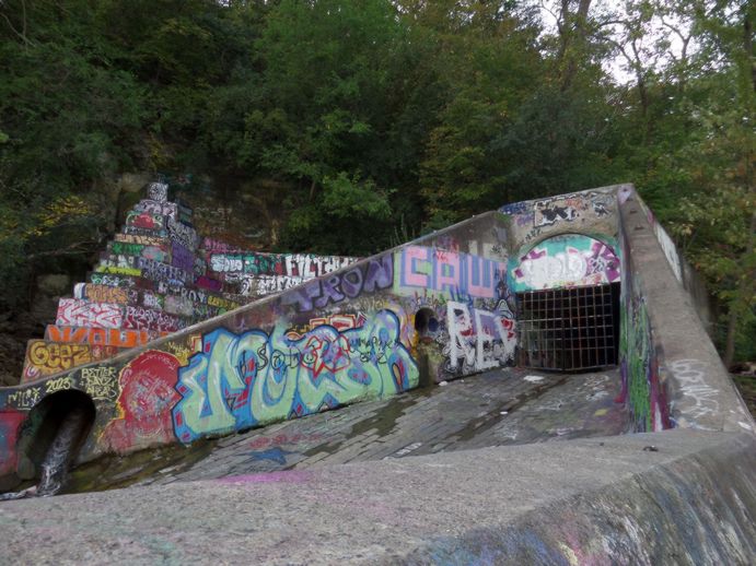 Big graffitied structure with steps too big for a person on the left and a metal gate on the right leading to a tunnel.