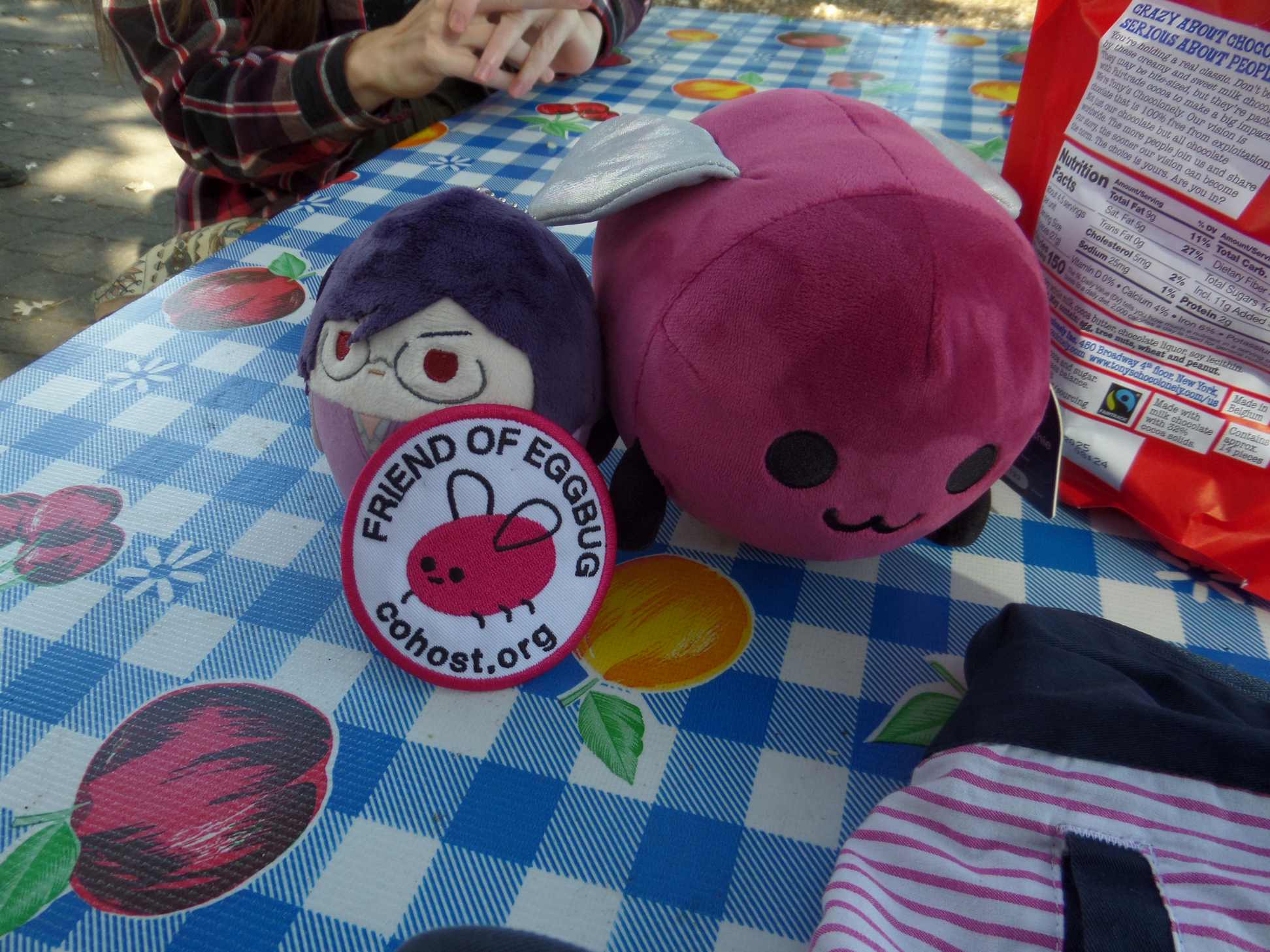 An Eggbug plush, a Gundam plush, and an Eggbug patch on a plaid picnic table cover.