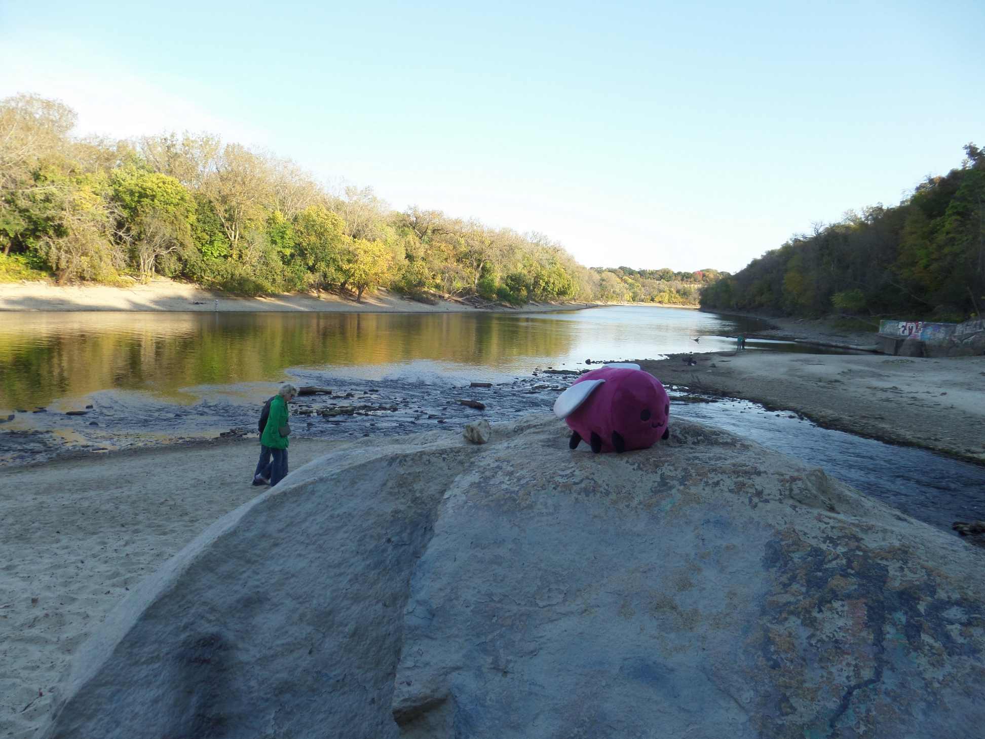 Eggbug plush on a large stone on a beach with a river in the background.