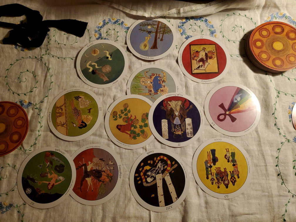 12 circular tarot cards are spread out roughly in a circle on top of an embroidered white cloth.