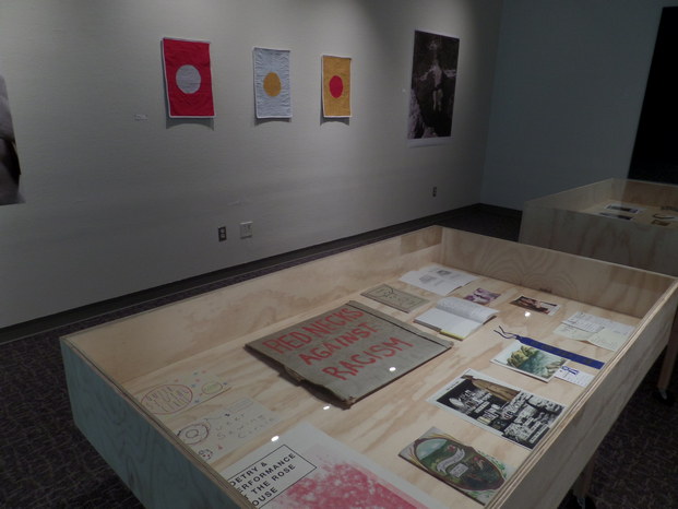 Queer ephemera in a gallery. Zines, art, and a yard protest sign that says 