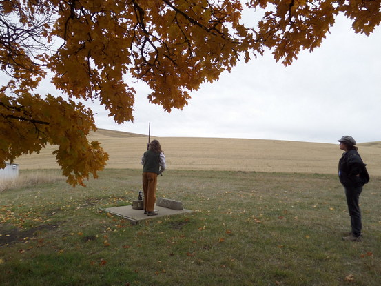 a person using an old water pump under some golden leaves. someone watches.